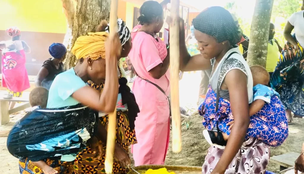 In Kailahun District, a group of mothers and caregivers preparing nutritious weaning foods for their babies using locally available ingredients, including rice or corn with legumes such as groundnuts and local vegetables.
